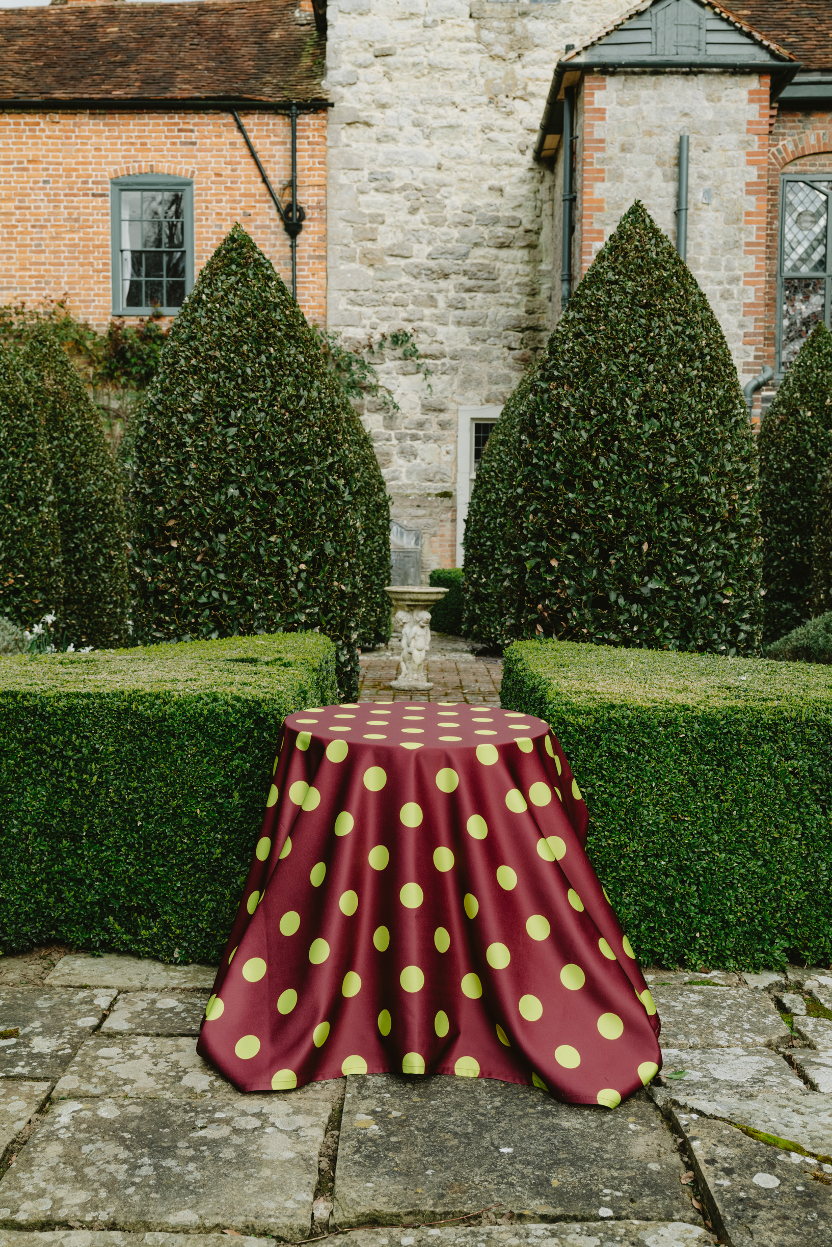 Image of a burgundy and lime green polka dot tablecloth on a table outside of a stone hosuse in a garden.
