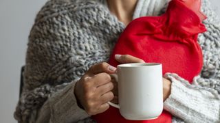 A woman in a thick grey cardigan cuddles a red hot water bottle and a white mug of hot tea