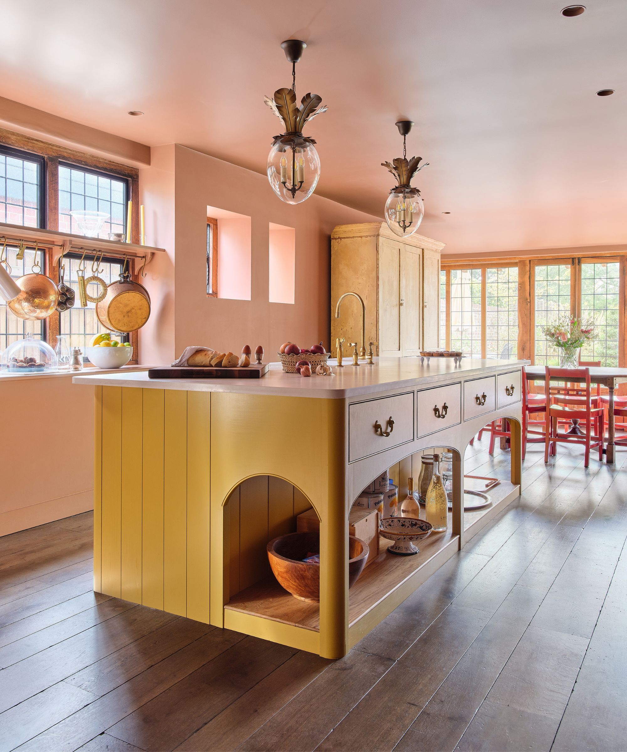 Character-filled kitchen with a mustard-yellow island, curved details, warm wood floors, vintage lighting, and soft pink walls for a cozy, eclectic feel.