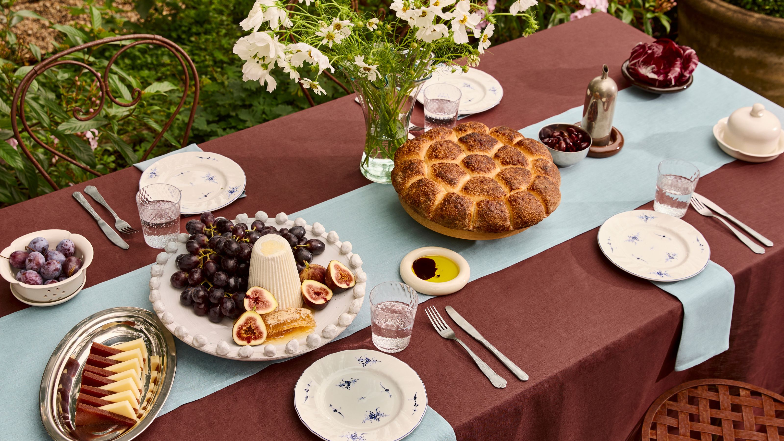 an outdoor table with a maroon tablecloth, blue table runner and napkins, set with bread, cheese and fruit
