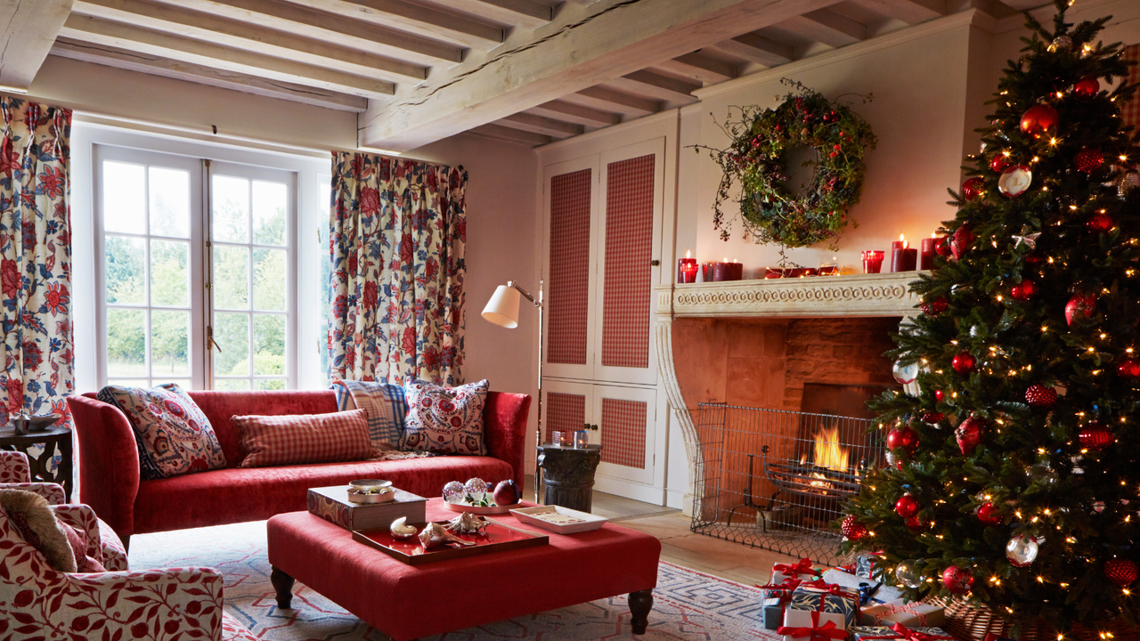Living room with lit fire and decorated Christmas tree with red furniture and red patterned soft furnishings. Cottage feel with a low ceiling and large open fireplace