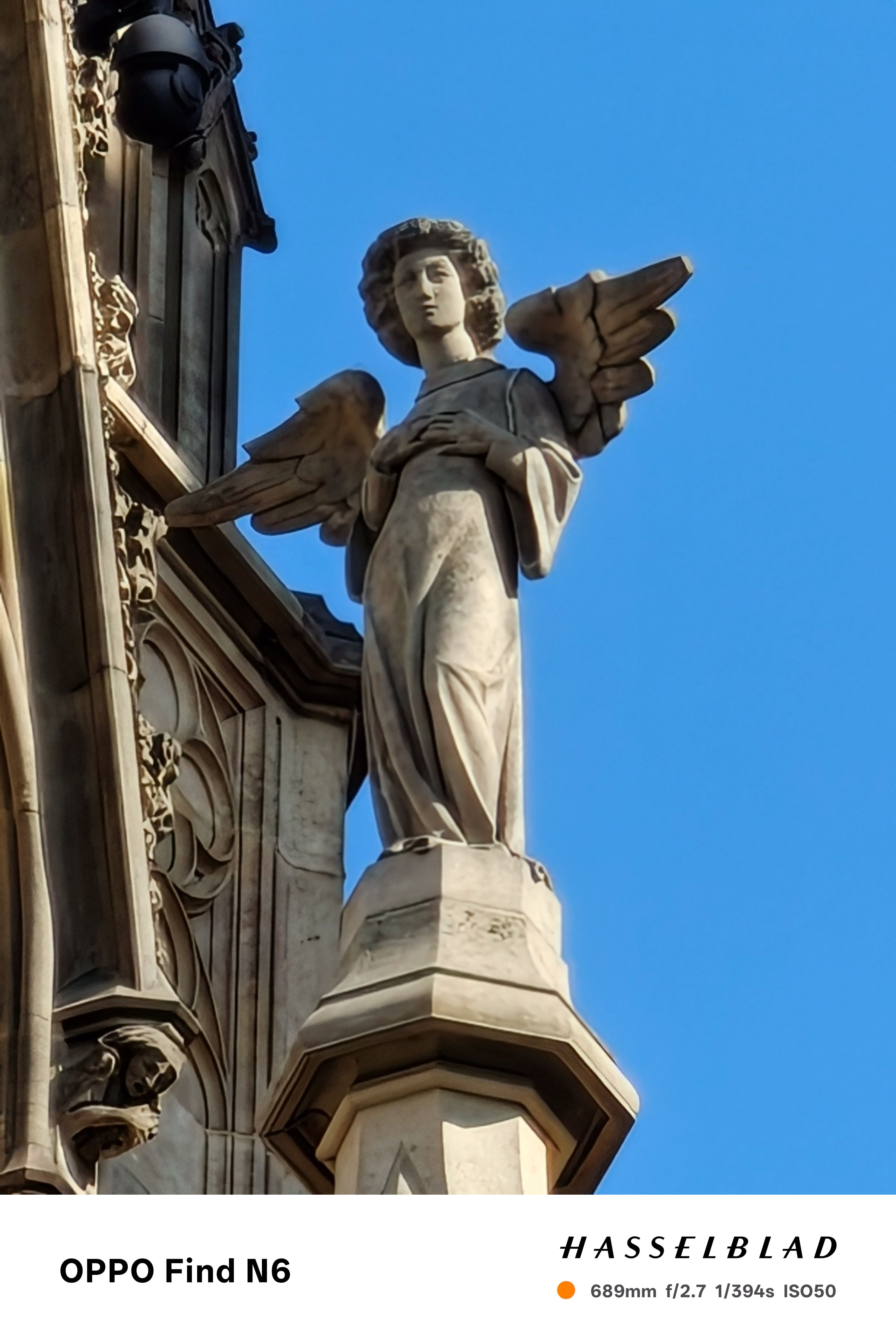 A high-zoom, close-up shot focusing on the same stone angel sculpture. The detail captured reveals the textures of the carved stone on the angel's face, hair, and flowing robes, showcasing the telephoto capabilities of the camera.