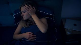 woman lying in bed in a dark room, holding a hand to her head as if in pain