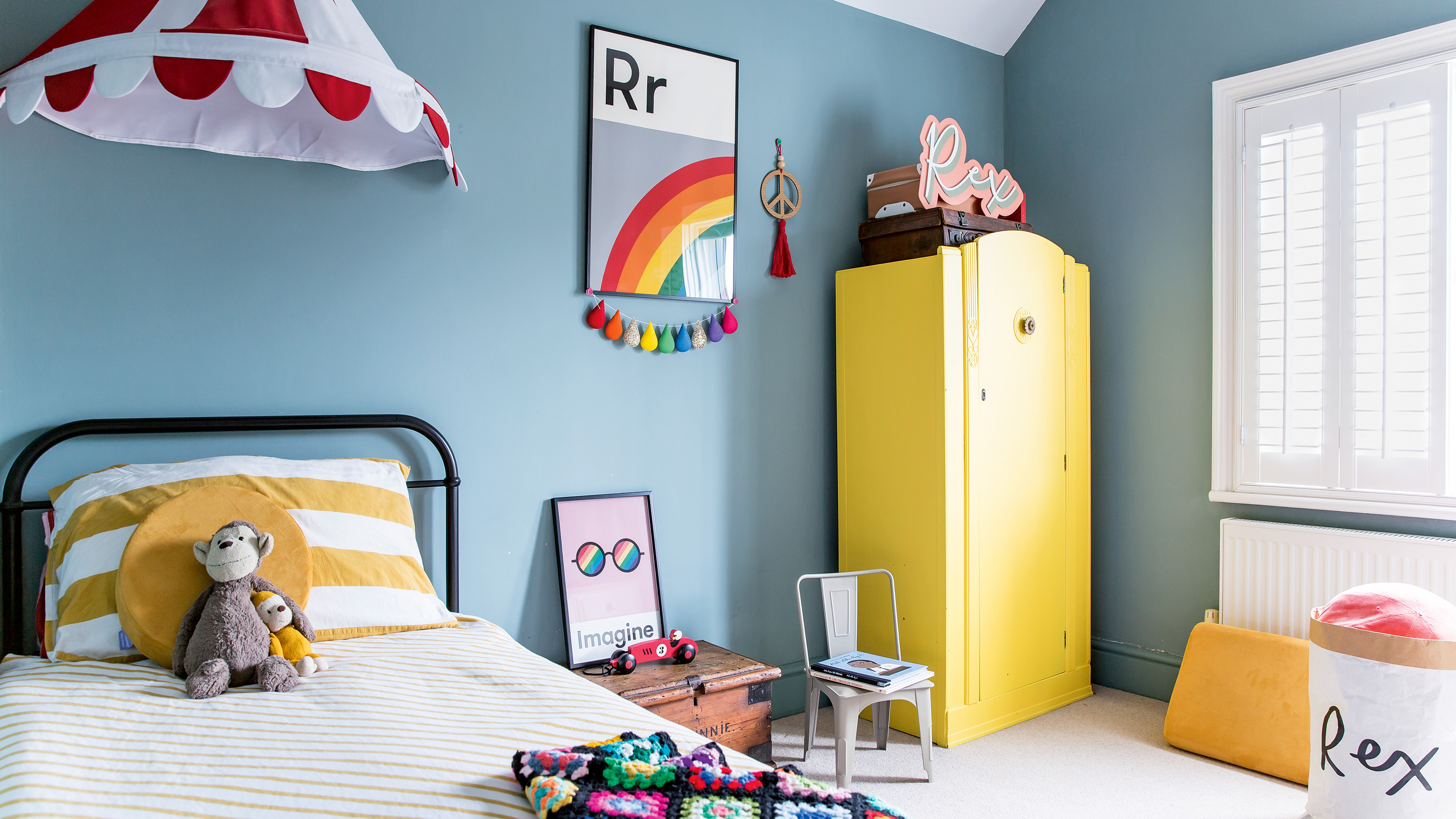 children's bedroom with blue walls, yellow wardrobe and cast iron bed with yellow and white striped bedlinen and two toy boxes lizzie orme