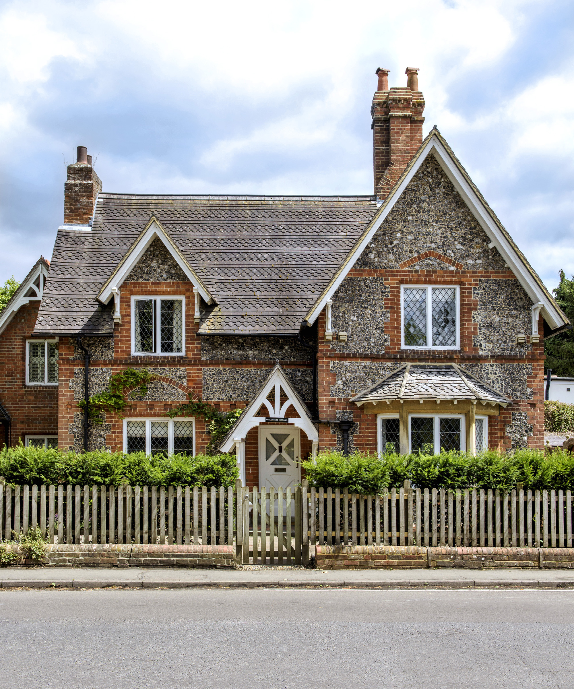 A small front garden with both a white fence and hedging, in front of a flint cottage.