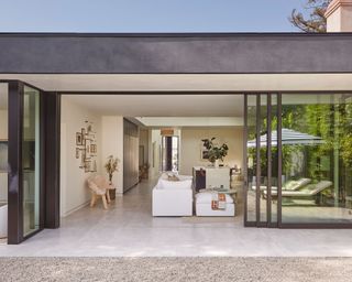 View from outdoors of the back of a modern property, large glass doors, looking into large open plan living area, stone gray flooring, black roof and white ceiling