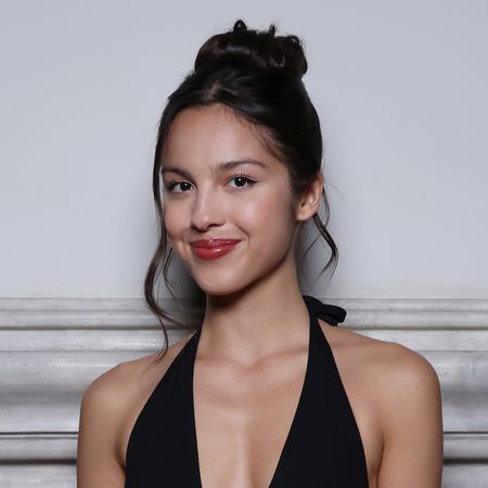 Olivia Rodrigo wears a top knot with pieces pulled out while standing in front of a white wall