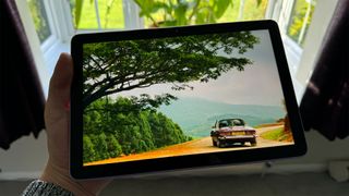 Amazon Fire HD 10 (2023) tablet held in hand in front of window with car and trees on screen