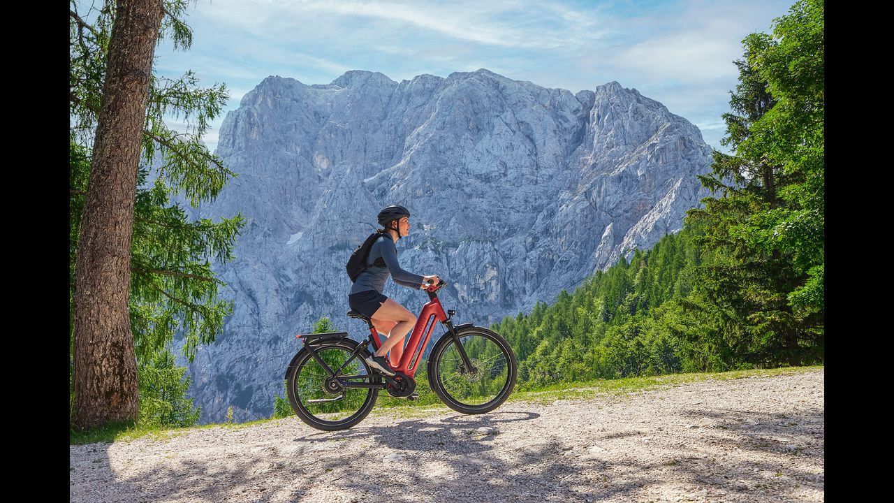 A woman riding an orange Eclipse ebike up a slight incline to the right with a mountain and evergreen trees in the background.