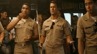 Glen Powell in uniform and holding a pool cue at a bar in Top Gun: Maverick. 