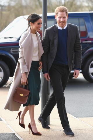 Meghan Markle wearing a green skirt, taupe coat and brown purse walking next to Prince Harry in front of a car