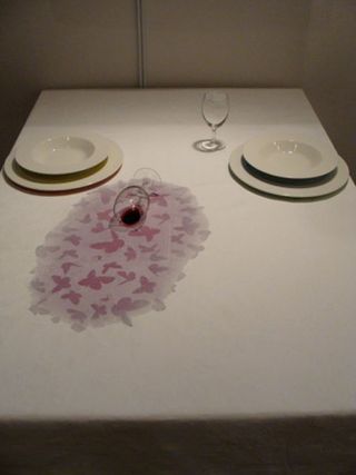 Dinner table dressed with a white table cloth and 2 stacked white plates on each side. One empty wine glass and one wine glass laying on the table with its content (red wine) spilled