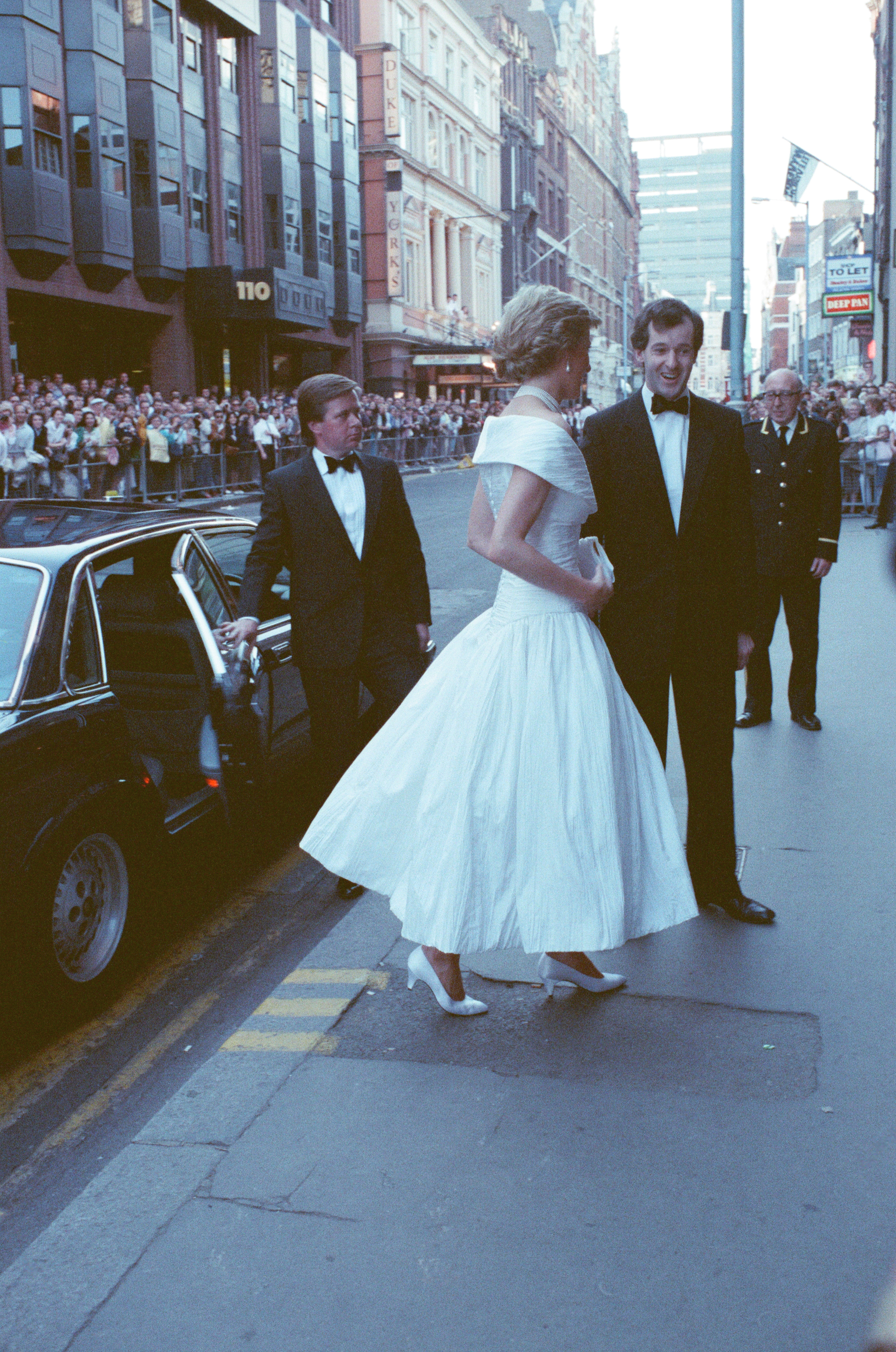 Princess Diana wearing a white dress with her bodyguard standing behind her and holding the car door