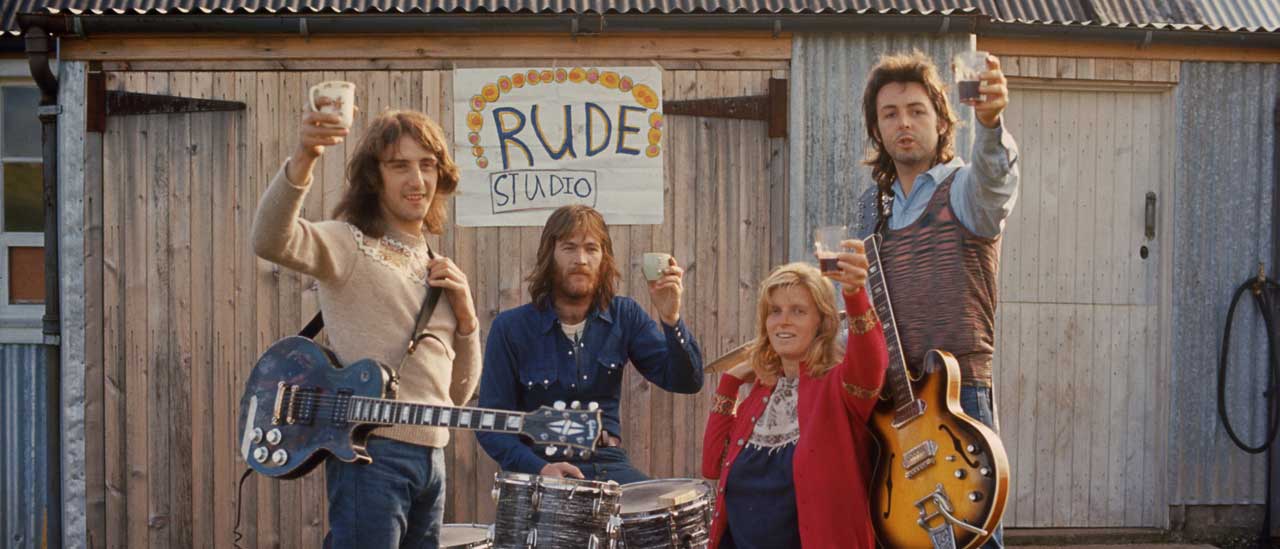 Wings with their instruments raising glasses to the camera outside a barn with a hand-painted sign 'Rude Studio'