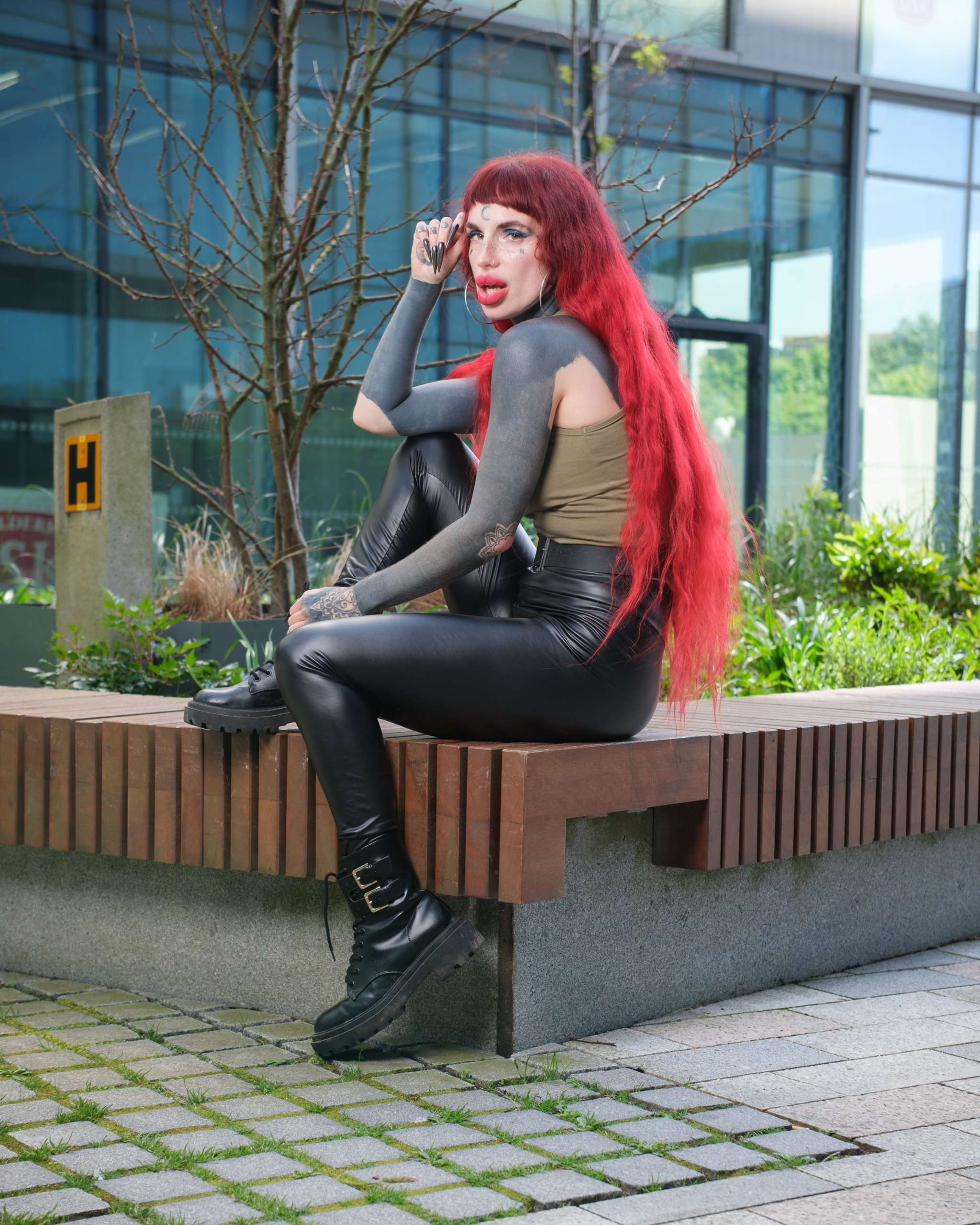 Red-haired person with dark body paint and long black nails posing outdoors in a belted olive dress against leafy greenery.