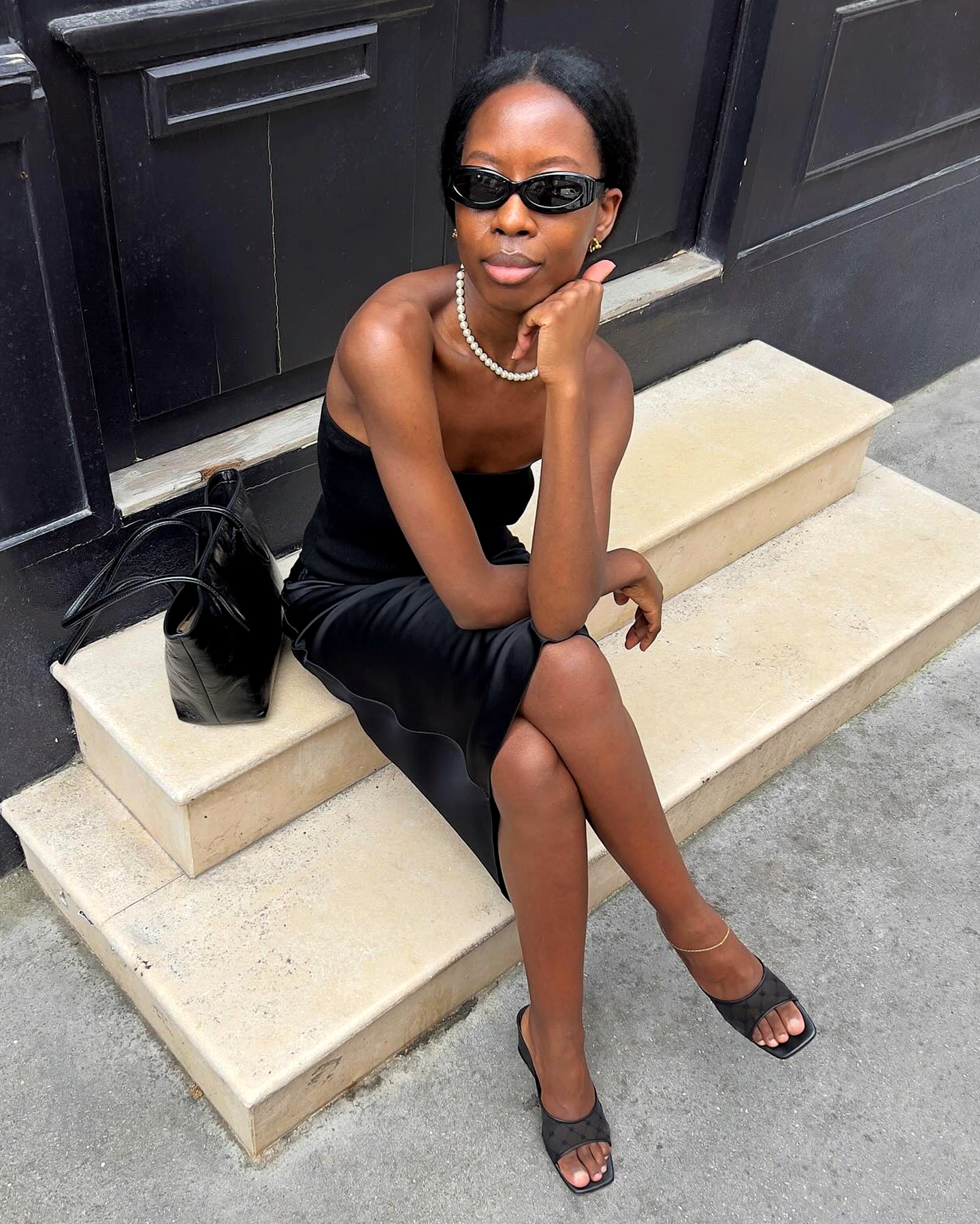 Style influencer Sylvie Mus poses on a Paris sidewalk wearing black oval sunglasses, a pearl necklace, a black sleeveless tube top, a black slip skirt, a black shoulder bag, and black mule heels.