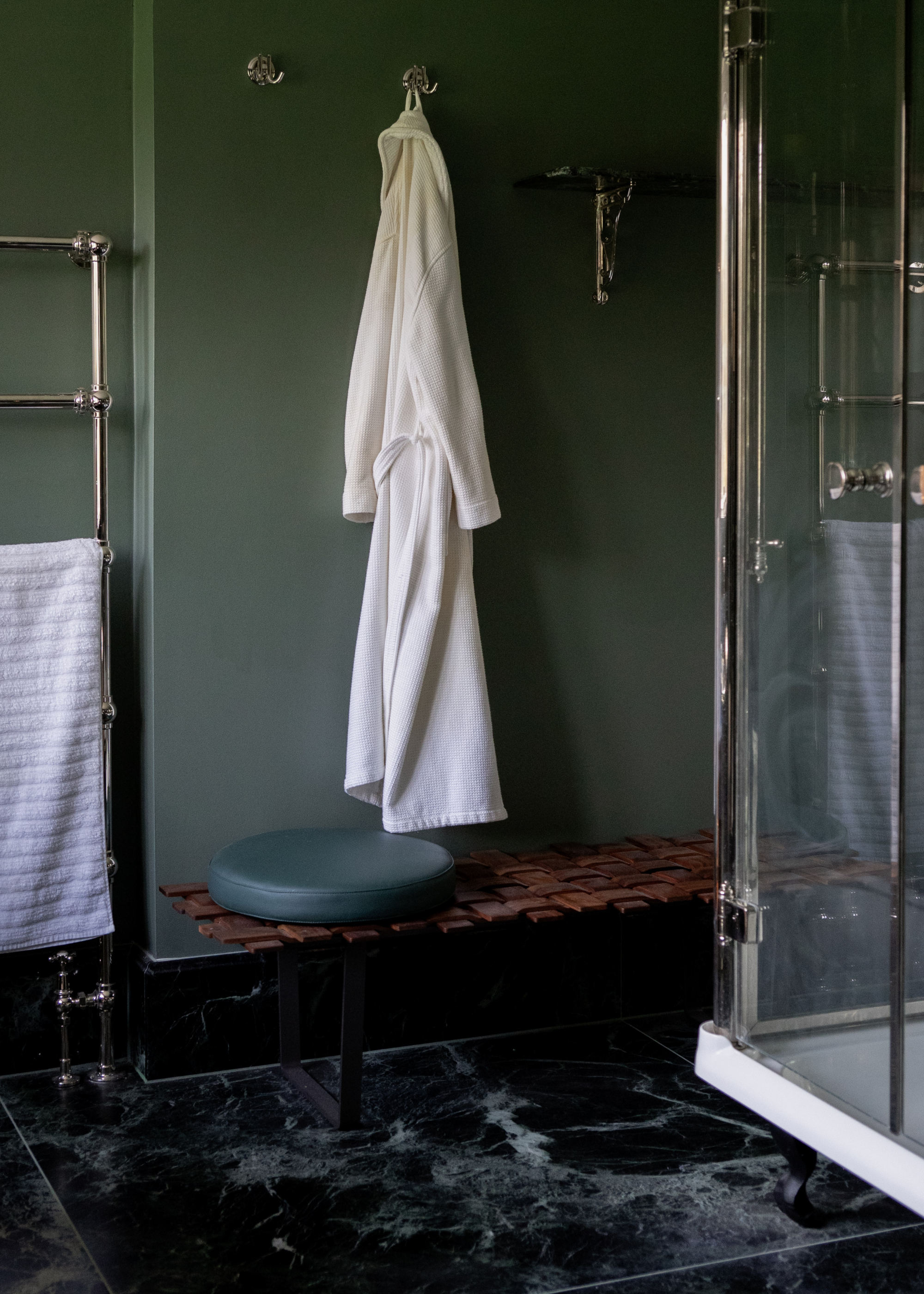A forest green bathroom with dark green marble floor tiles, a shower to the right and a woven wood effect bench with a matching green seat cushion, and a white dressing gown hung above on chrome hooks.
