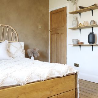 A bedroom with a plaster-effect brown-painted wall, a rattan headboard and floating shelves displaying decorating objects made with ceramic and glass