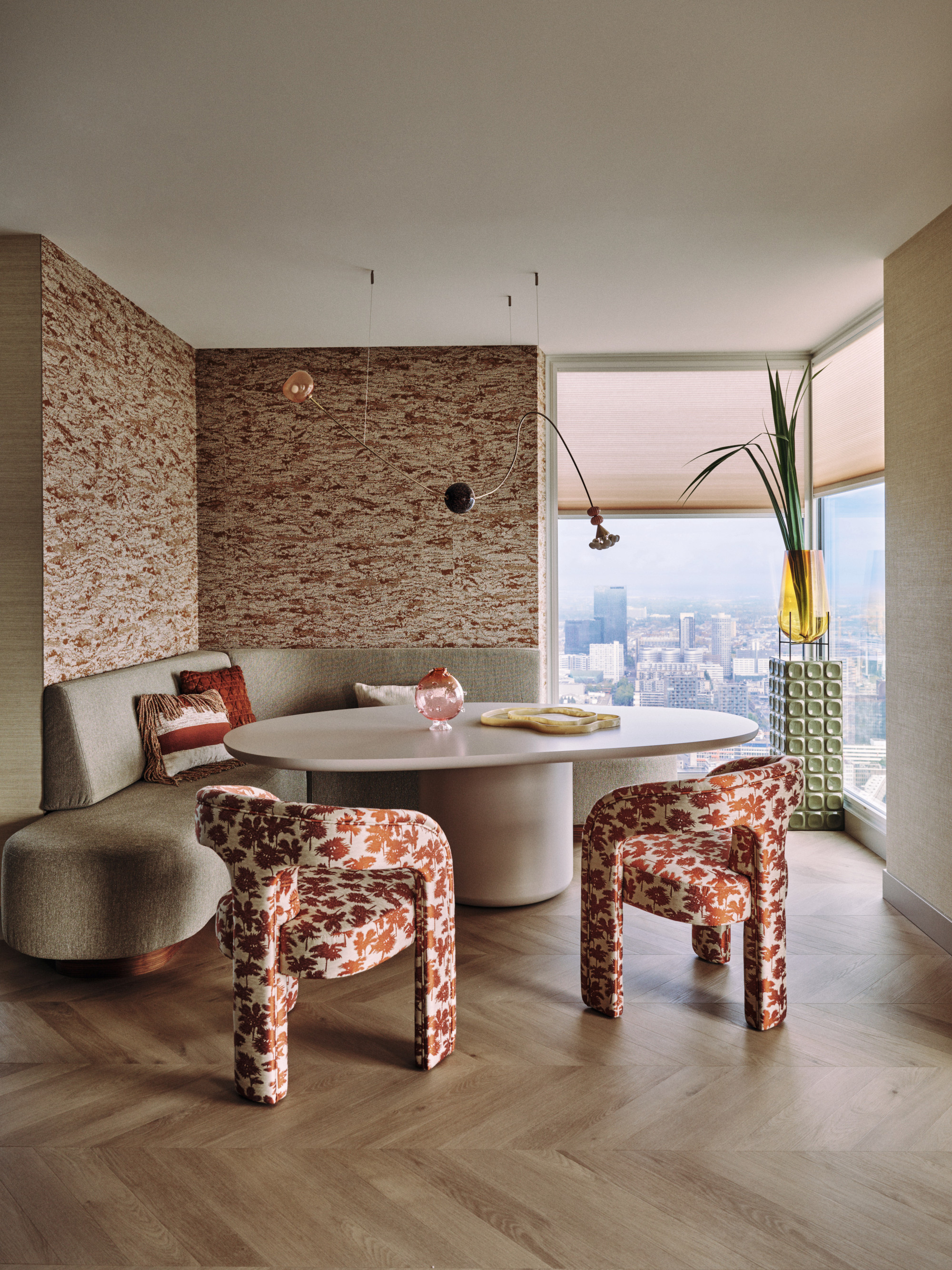 Dining nook with corner banquette, oval table, patterned armchairs, patterned wallpaper and chevron flooring