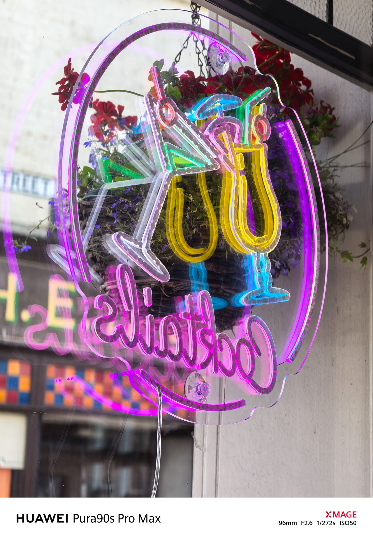 Bright multicolored neon cocktail sign displayed in a window with flowers behind it.