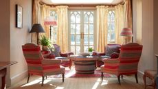 A maximalist seating area with patterned red chairs surrounding a stone coffee table. Yellow drapes cover floor-to-ceiling windows and patio doors behind. 