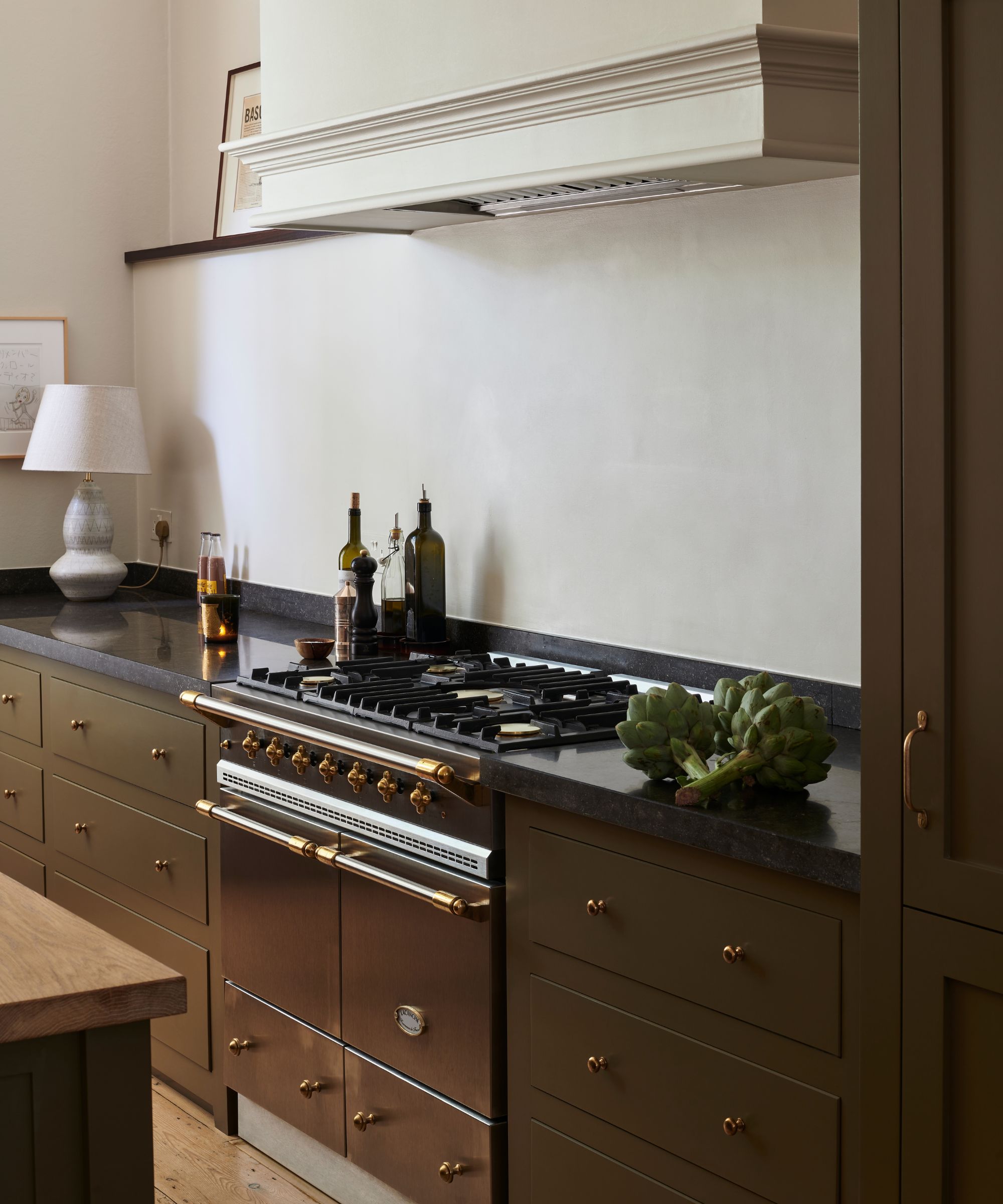 Kitchen detail showing a professional copper and black range stove, dark countertops, olive-green drawers with brass knobs, and fresh artichokes
