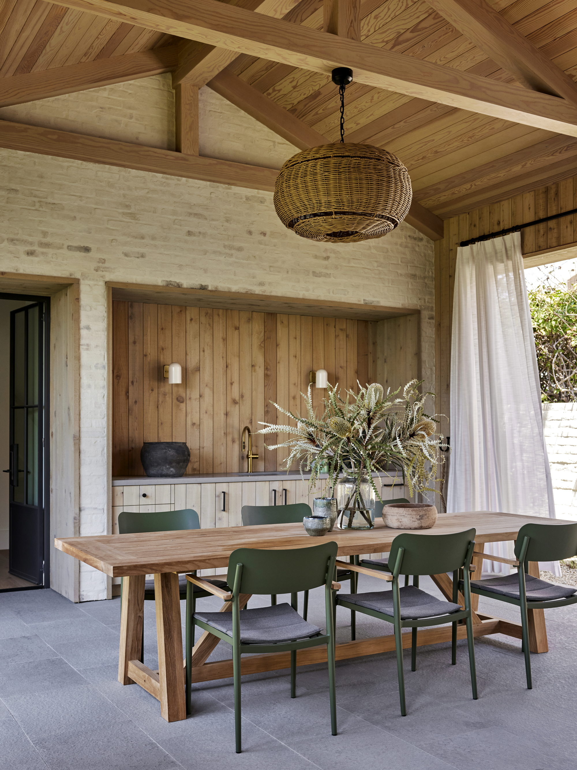 Image of a wooden outdoor dining table underneath a structure. There are six green dining chairs around it and a rattan pendant light overhead.