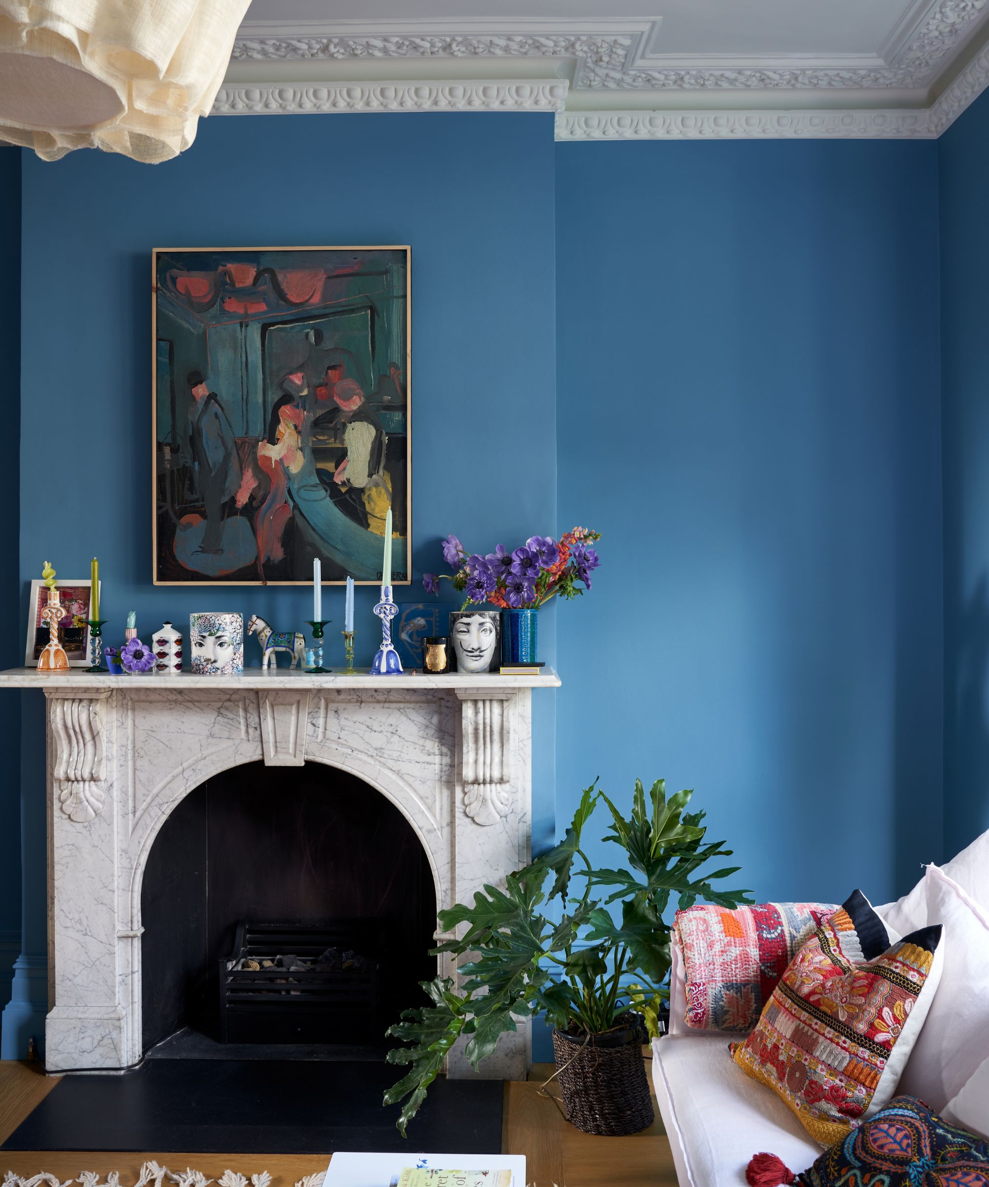 Living room painted in Farrow &amp;amp; Ball's Bothy Blue, a marble-white fireplace with candlesticks and decor on top, an impressionist painting above it, a white sofa, and colourful embroidered cushions.