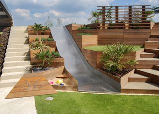 A slide, sandpit and zig-zag wooden steps integrated into sloped backyard ideas.