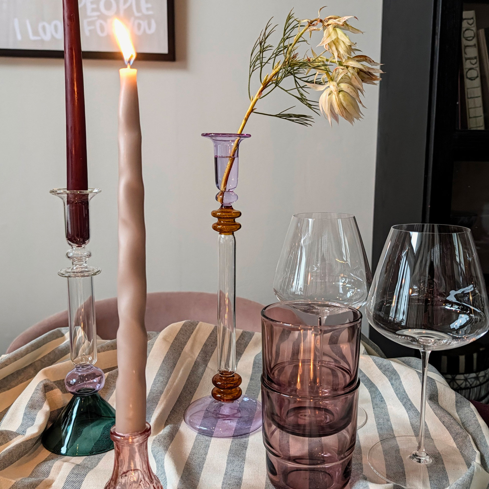 Three coloured glass candlestick holders beside wine and drinking glasses on striped tablecloth