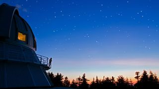 A person stands on the balcony of an observatory and gazes out to a star filled sky with trees silhouetted below