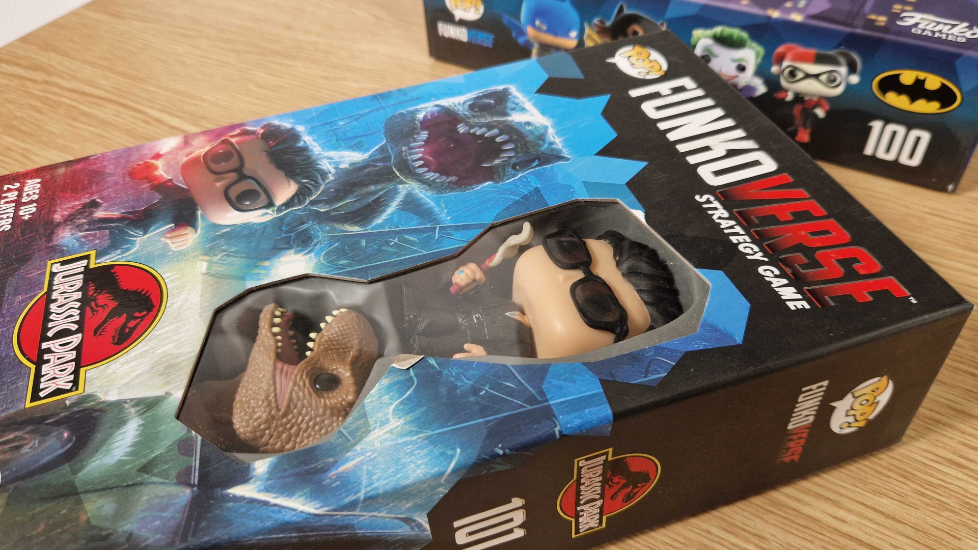 Funkoverse Jurassic Park and DC box set lying on a wooden surface beside one another