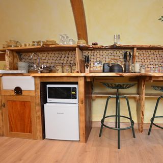 a hobbit house wooden kitchen and dining area, one long bench all along a yellow wall, with green stools under the dining bench and an earthy mozaic tile splashback