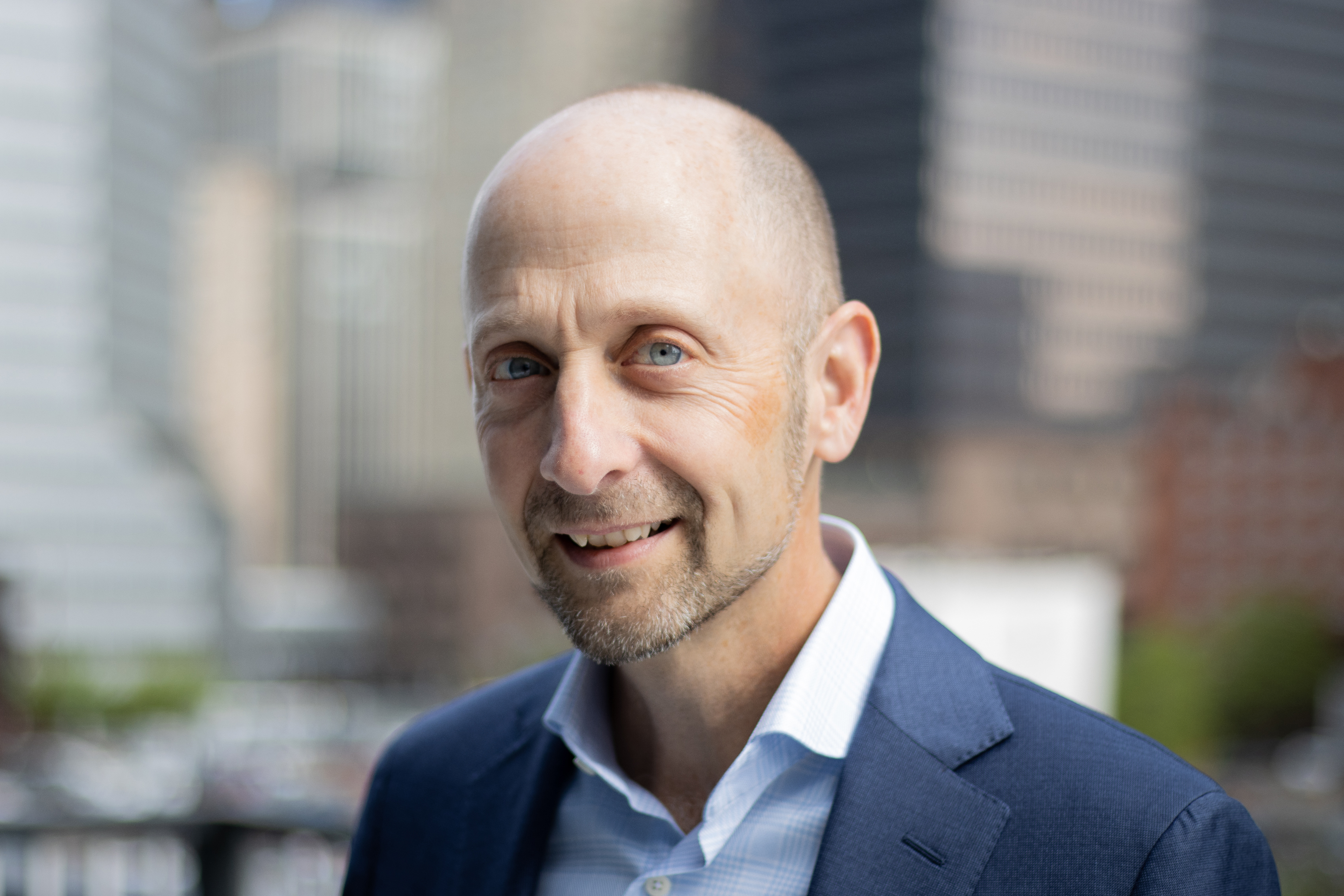 An image of a bald man in a blue jacket and shirt against a blurred background. The man is a doctor.