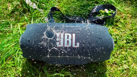 the jbl xtreme 5 with water splashing on it outside in the park showing its waterproofing