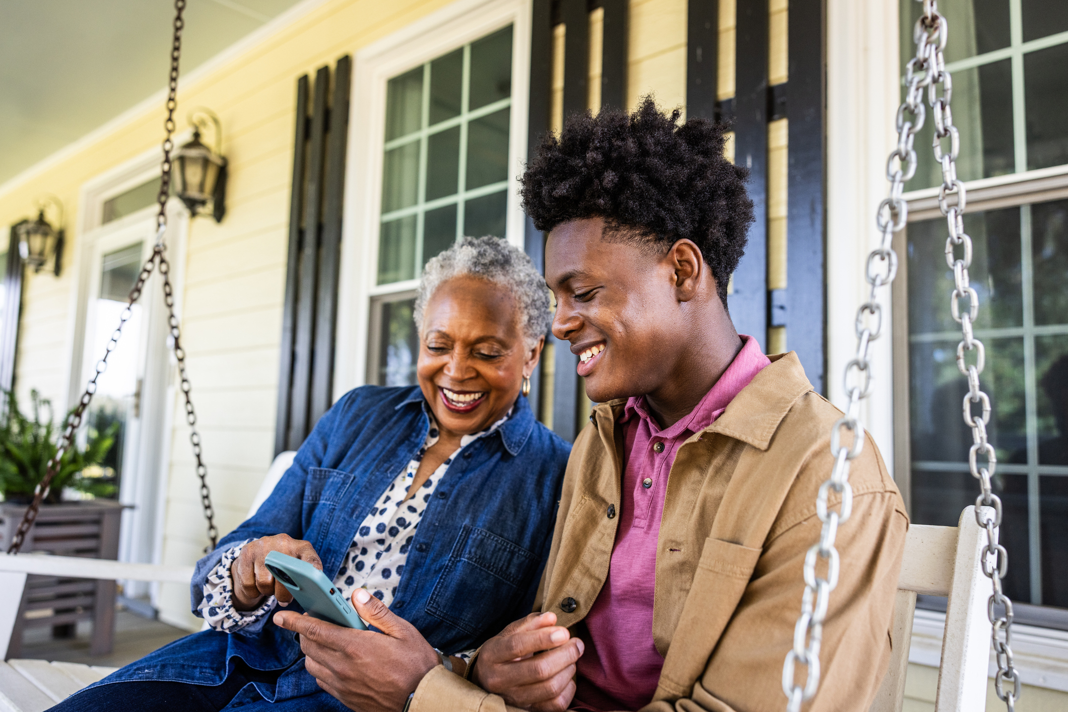 A grandmother and grandson sit on a porch swing looking at pictures on a cell phone