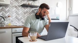 Stressed man in a kitchen sat in front of a laptop with a coffee