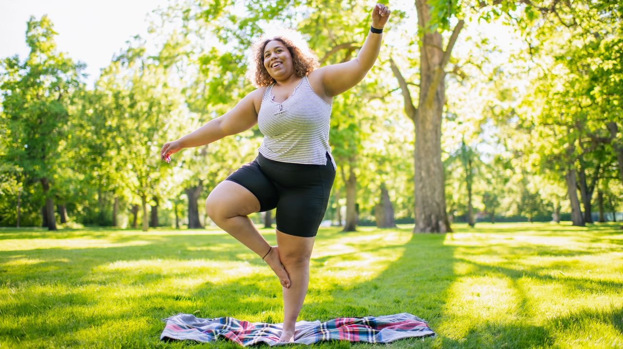 A woman balances on one leg in a park on a blanket. She holds one arm out to the side and one arm above her head. Behind her we see lots of green, healthy trees and dappled sunlight.