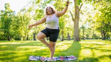 A woman balances on one leg in a park on a blanket. She holds one arm out to the side and one arm above her head. Behind her we see lots of green, healthy trees and dappled sunlight.