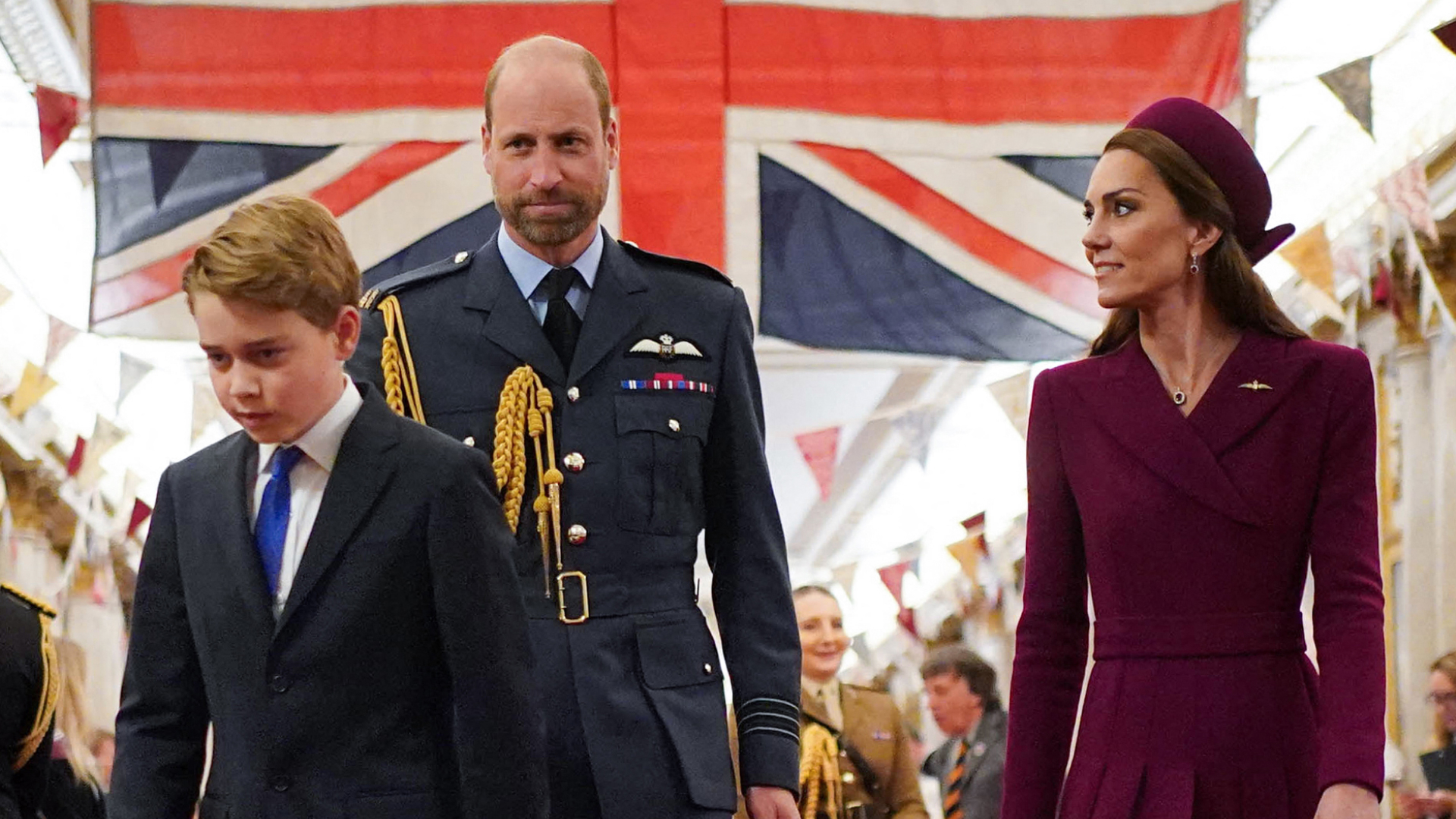Prince George wears a navy suit and walks in front of a large Union Jack flag with his parents, Prince William in a navy military uniform and Princess Kate in a dark red coat dress