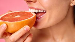 Woman eating a half grapefruit smiling against pink background
