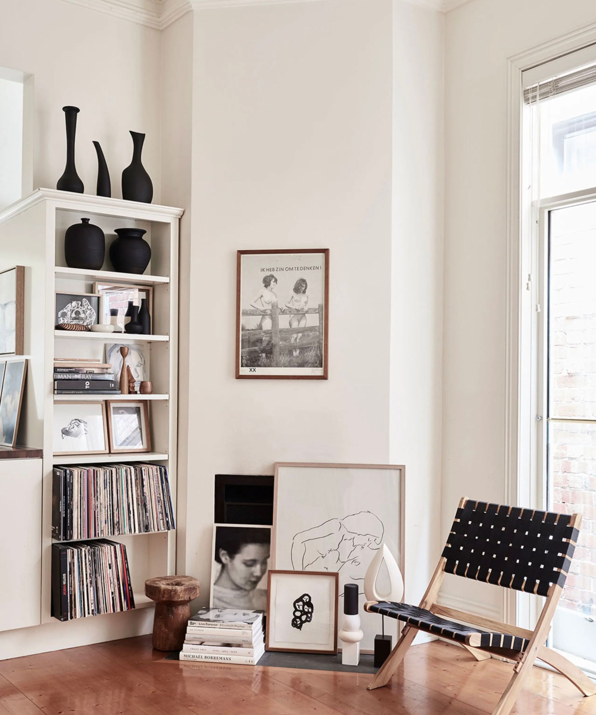 Modern living room with white walls, prints on the floor, a woven lounge chair, and a bookcase filled with records, vases and photographs