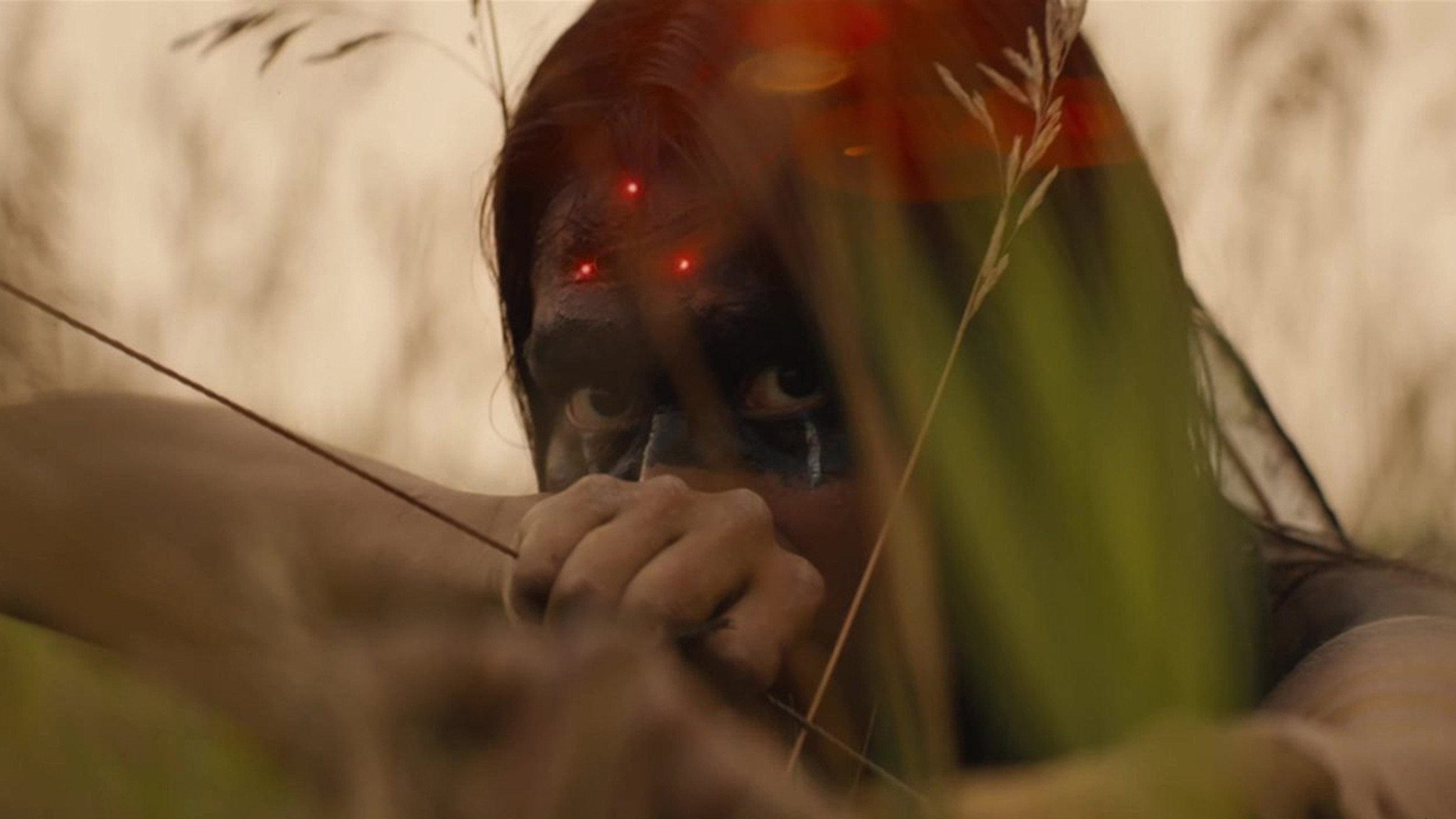 A close-up of Naru crouching amongst tall grass and pulling back the string of a bow. There are three laser points sitting on her forehead.