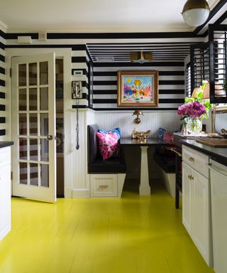 black and white kitchen with striped wallpaper, banquette seating, bright yellow painted floorboards, artwork, white cabinetry, black countertops