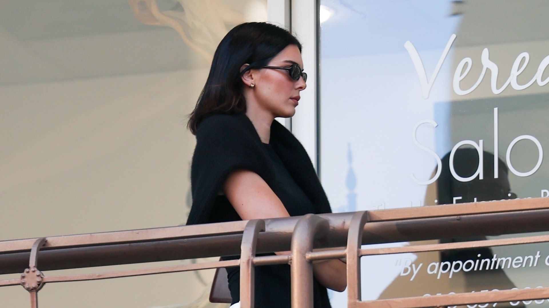 Kendall Jenner walks outside sushi park wearing a black top with a black jumper knotted around her shoulders and cream satin trousers.