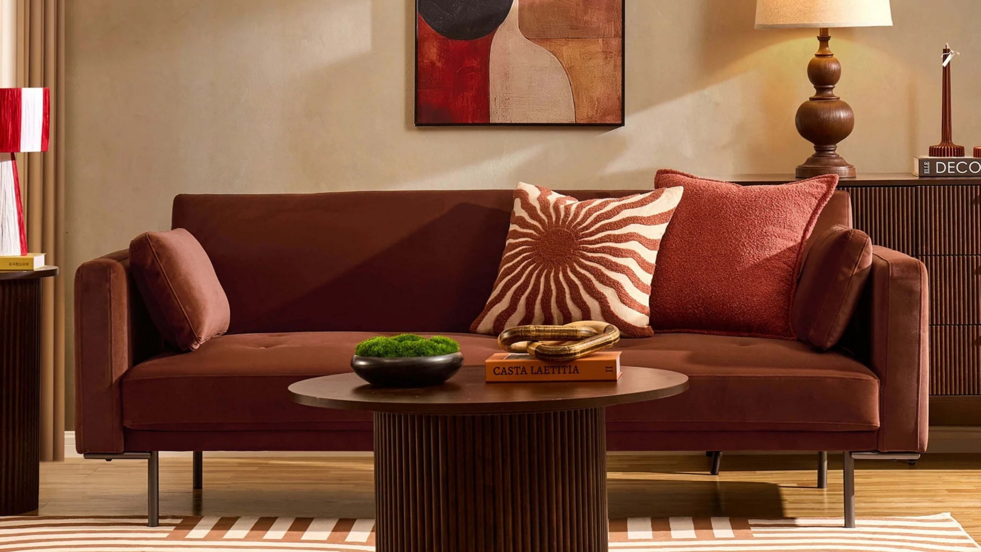 Image of a brown, mid-century modern sofa bed in a modern living room. There are two throw pillow on the sofa and a round, wooden fluted coffee table in front of the sofa.