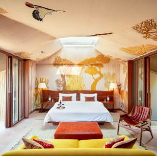 RAAS Chhatrasagar hotel room, neutral floor and scatter rugs, white dressed double bed with orange cushions and bottle of red wine and quarter filled glasses on a gold tray, brown ottoman at the foot of the bed, wooden frame glass doors on the left and right walls, yellow settee across from the bed, rustic colour artwork of trees, plants, deer and birds illustrated onto the walls and ceiling, red leather chair to the right, floor standing lamps lit up either side of the bed