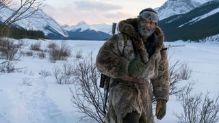Hold the Dark image of a man walking in a thick fur coat looking away from the camera and surrounded by trees, mountians and snow. 