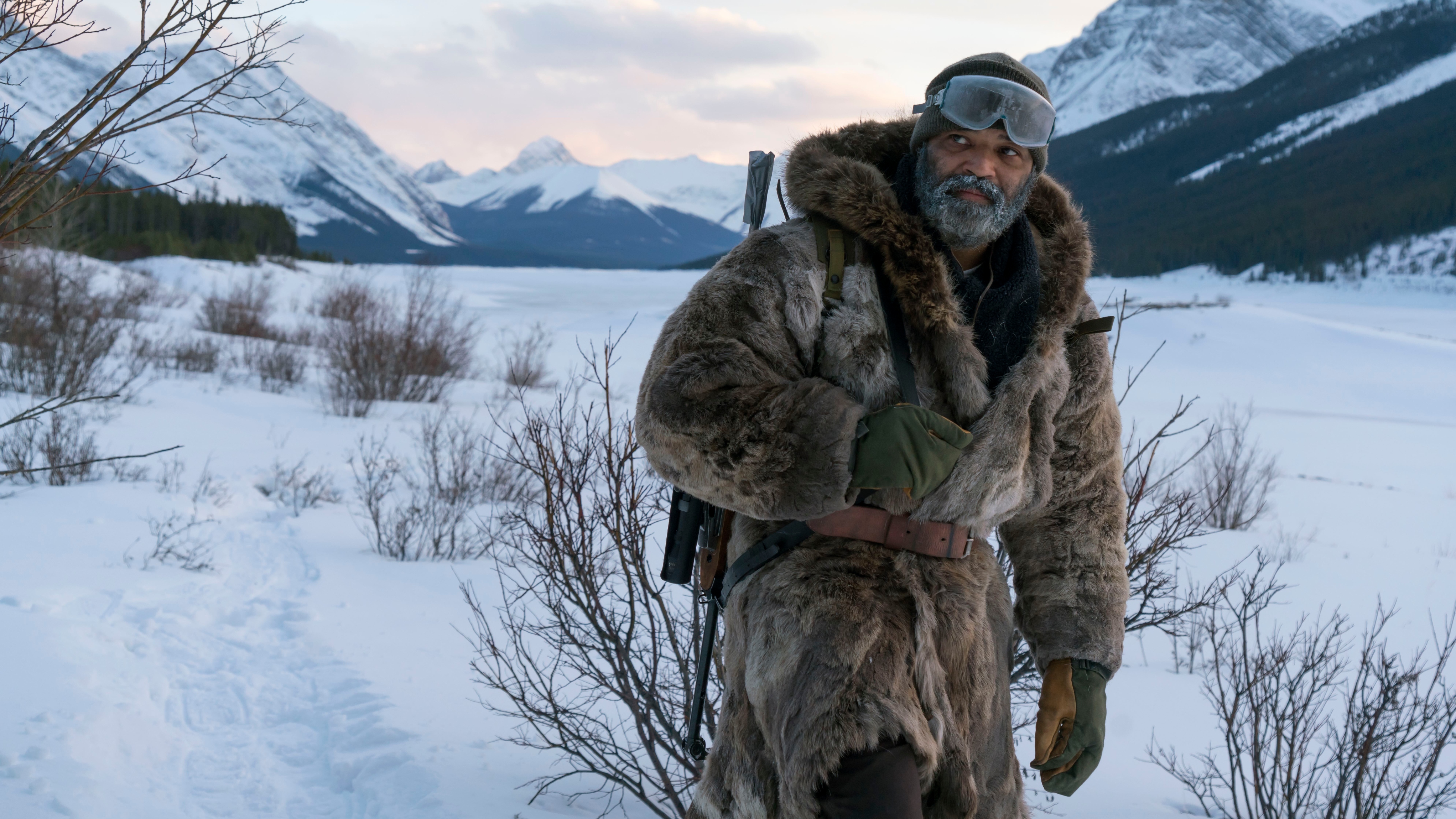 Hold the Dark image of a man walking in a thick fur coat looking away from the camera and surrounded by trees, mountians and snow.