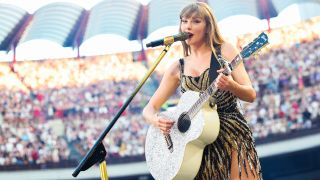 Taylor Swift in a black and gold dress, playing a silver guitar and singing into a mic on July 13, 2024 in Milan, Italy.